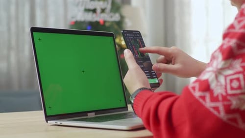 Male Using The Smartphone Checking The Stock Market While Working On Green Screen Laptop