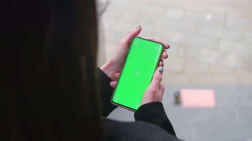 Close Up Female Hands Holding a Vertical Mobile Phone with Green Screen Against Background of Urban
