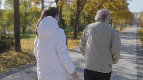 Back View of Elderly Man with Nurse Strolling in Serene Autumn Park