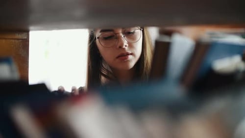 Blick durch die Regale einer Studentin mit Brille, die in der Bibliothek nach Büchern sucht