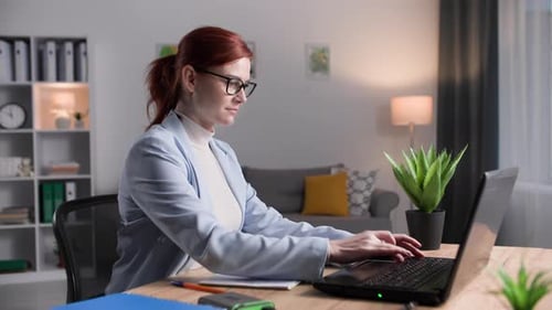 Junge Frau mit Brille macht aus der Ferne Arbeit und macht Notizen im Notizbuch, während sie am Laptop im Zimmer sitzt