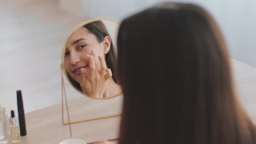 Woman Applies Lotion to Face in Mirror