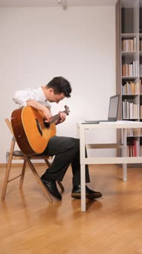 Vertical Video Man Holding an Acoustic Guitar and Looking at a Laptop Screen Learning to Play Music