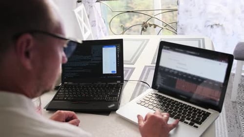 Fokussierter IT-Spezialist für Brillen schreibt Programmcode auf zwei Laptops, während er zu Hause arbeitet