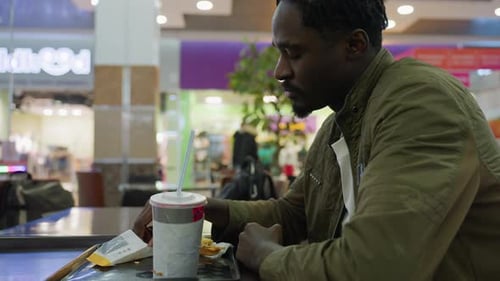 Young Man Sitting Alone Eating Fries At Table In Food Court
