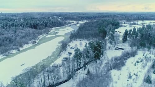Aerial drone panning shot over cottages beside frozen river and forest along winter landscape on a c