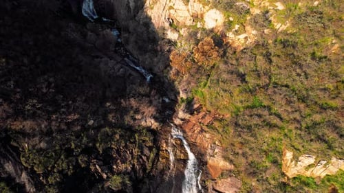 Luftaufnahme eines kaskadierenden Wasserfalls, umgeben von felsigem Gelände und Vegetation in einem bergigen Gebiet