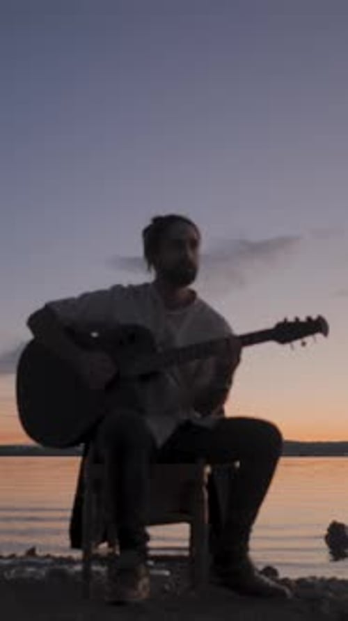 Silhouette of a Man Playing Guitar Sitting on a Chair By the Lake on Colourful Sunset Background