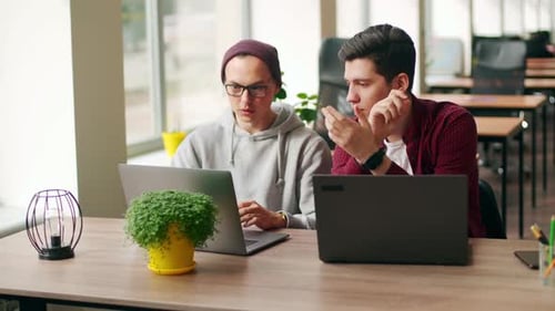 Dos compañeros de trabajo hipster comparten ideas sobre un proyecto sentados con computadoras portátiles en una espaciosa oficina de espacio abierto