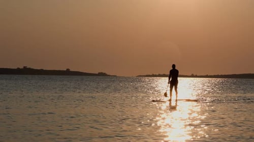 Man is Paddling a Surfboard on a Calm Lake at Sunset