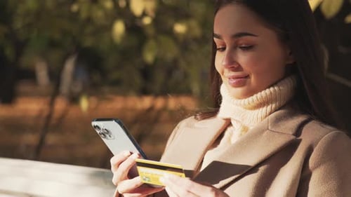 Young Woman Walks in Autumn Park in Nature with Phone and Bank Card in Hands and Pays for Purchases