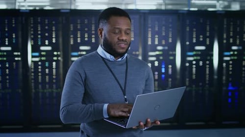Black Data Center IT Technician Standing in the Middle of a Server Rack Corridor with a Laptop Comp