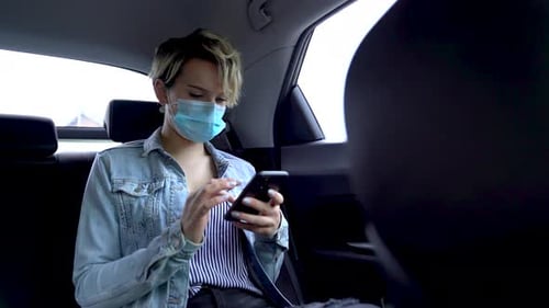 Girl In Mask Sits In A Car The Back Seat Looks Into SmartphoneWide shot. Coronavirus