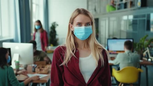 A Young Woman in an Office Setting is Wearing a Mask While Surrounded By Her Colleagues