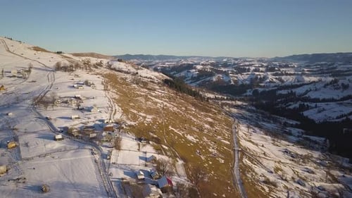 Aerial View of Small Village with Scattered Houses on Snow Covered Hills in Winter and Bare Dark