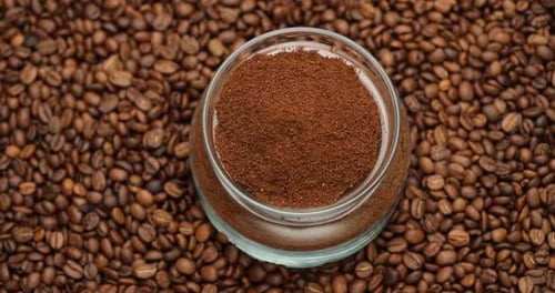 Glasschale voller gemahlener Kaffee auf einem rotierenden Hintergrund aus frisch gerösteten Kaffeebohnen