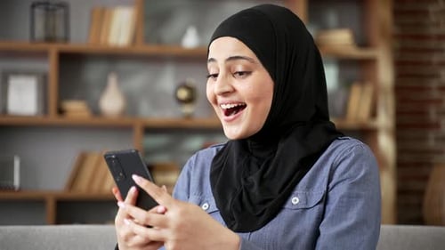 Happy excited young woman in hijab celebrating success by reading great news on smartphone sitting