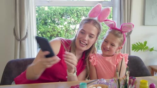 Mother extending phone, daughter wearing bunny ears, sharing pictures while crafting eggs at home
