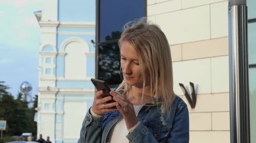 Portrait blonde young woman walking streets of city and using smartphone