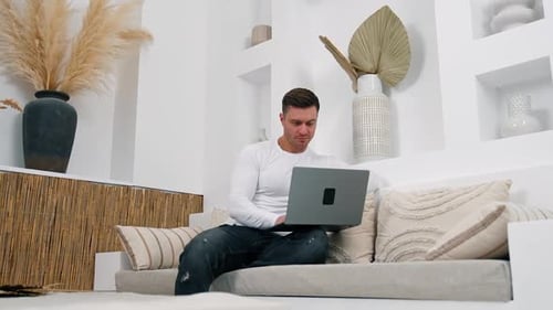 Dark-haired Caucasian male sits on sofa focused on laptop. Working remote from home. Low angle view.