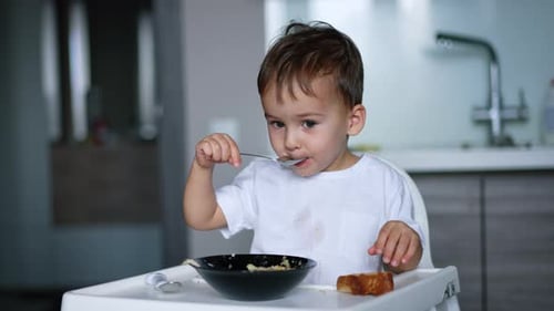 Menino fofo em camiseta branca sentado em uma cadeira alta na cozinha.