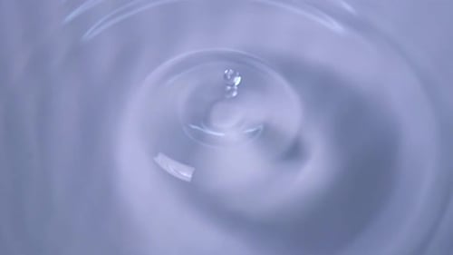 A Drop of Water Falls Forming Circles on the Surface of the Water Against a Pale Blue Background