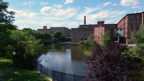 Drone shot slowly flying forward through a few trees in a park toward a river and an old brick mill