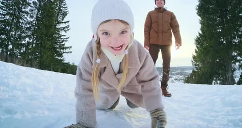 Family enjoy winter time. Happy cute girl crawls on the snowy field with her parents on the backgrou