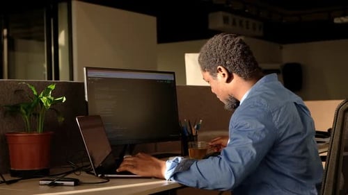 Homem negro escrevendo linhas de código em um PC de mesa com dois monitores e um laptop ao lado em um escritório elegante