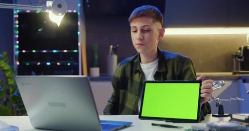 Adolescente concentrado sentado en el escritorio hace una videollamada, conferencia, discurso y muestra una tableta con pantalla verde