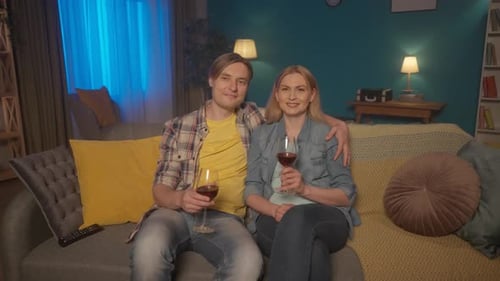 A Young Couple Sits Hugging on the Couch with Glasses of Red Wine and Watching TV A Man and a Woman
