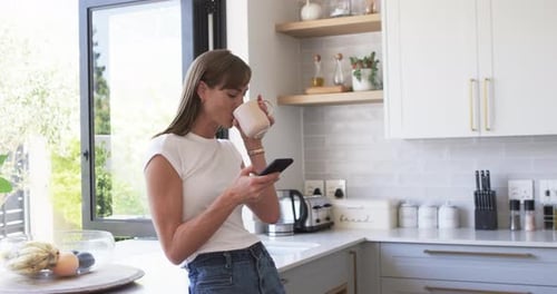 Woman Drinking Coffee and Using Phone in Kitchen
