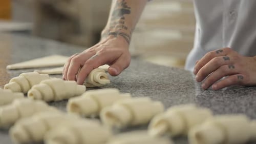 A baker forms a croissant from puff pastry at a bread factory. Close-up of male hands with tattoos.