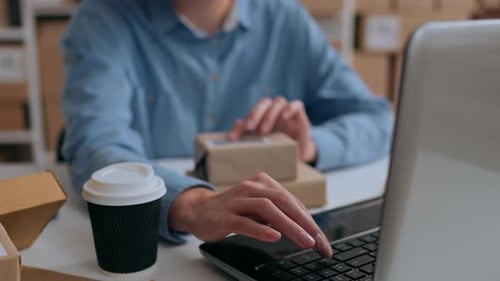 A Girl Works At A Laptop Laptop While Sitting In The Office Against The Background Of Boxes Of Goods