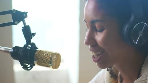 De perto, jovem afro-americana fala no microfone, senta-se à mesa na sala