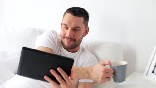 Man using tablet and drinking coffee in bed