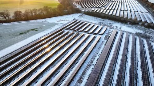 Solar farm in winter with rows of photovoltaic panels partially covered in snow, set against backdr