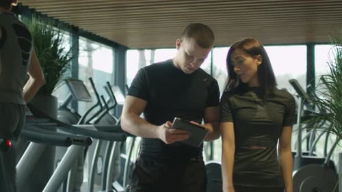 Athletic man and woman are using a tablet next to treadmills in a sport gym.
