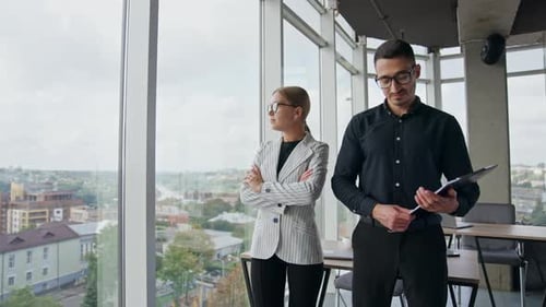Male and female colleagues standing in office near the panoramic window.
