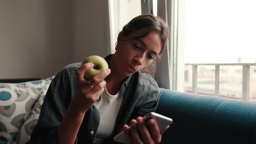 Young Adult Woman Eating Apple While Using Cellphone