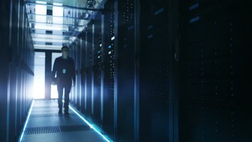 Un ingeniero de TI sostiene un portátil y recorre un centro de datos lleno de servidores en rack en funcionamiento.