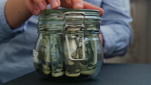 Female Hands Turn Glass Jar Full of American Currency Dollars Cash Banknote with Text 2027 Year