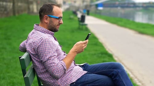 Young Man Texting on Smartphone Sitting on Bench in City 30s