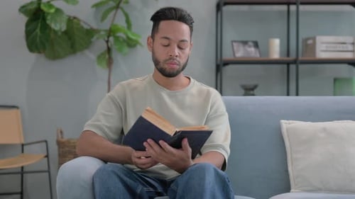 Hispanic Man Reading Book while Sitting on Sofa at Home