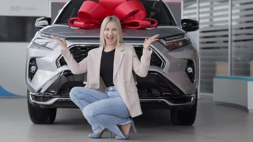 Happy business woman smiling and crouching next to new car with red bow in dealership