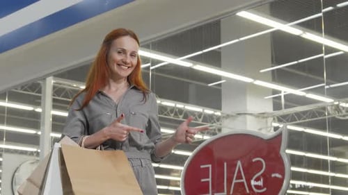 Charming Woman Joyfully Pointing at a Vibrant Sale Sign in a Bustling Store