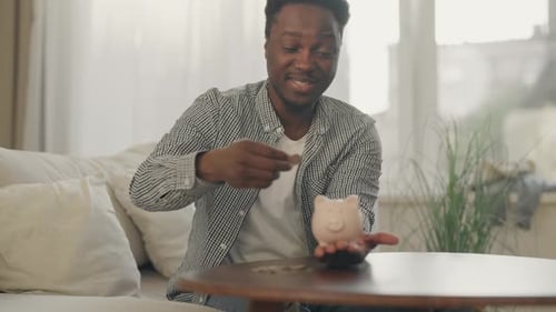 Porträt eines afrikanischen Mannes, der Geld in einer Münzbox spart und Münzen in ein Sparschwein steckt, junger erwachsener Mann