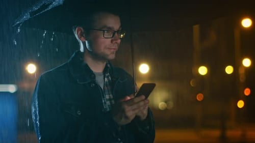 Young Caucasian Man in Glasses, Wearing a Jeans Coat and Square Shirt is Using a Smartphone Under a