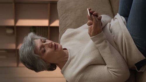 Vertical Screen Senior Woman Uses Smartphone While Relaxing on Couch at Home