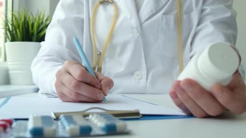 Female Doctor Holding White Pill Bottle and Writing Prescription in Clinic